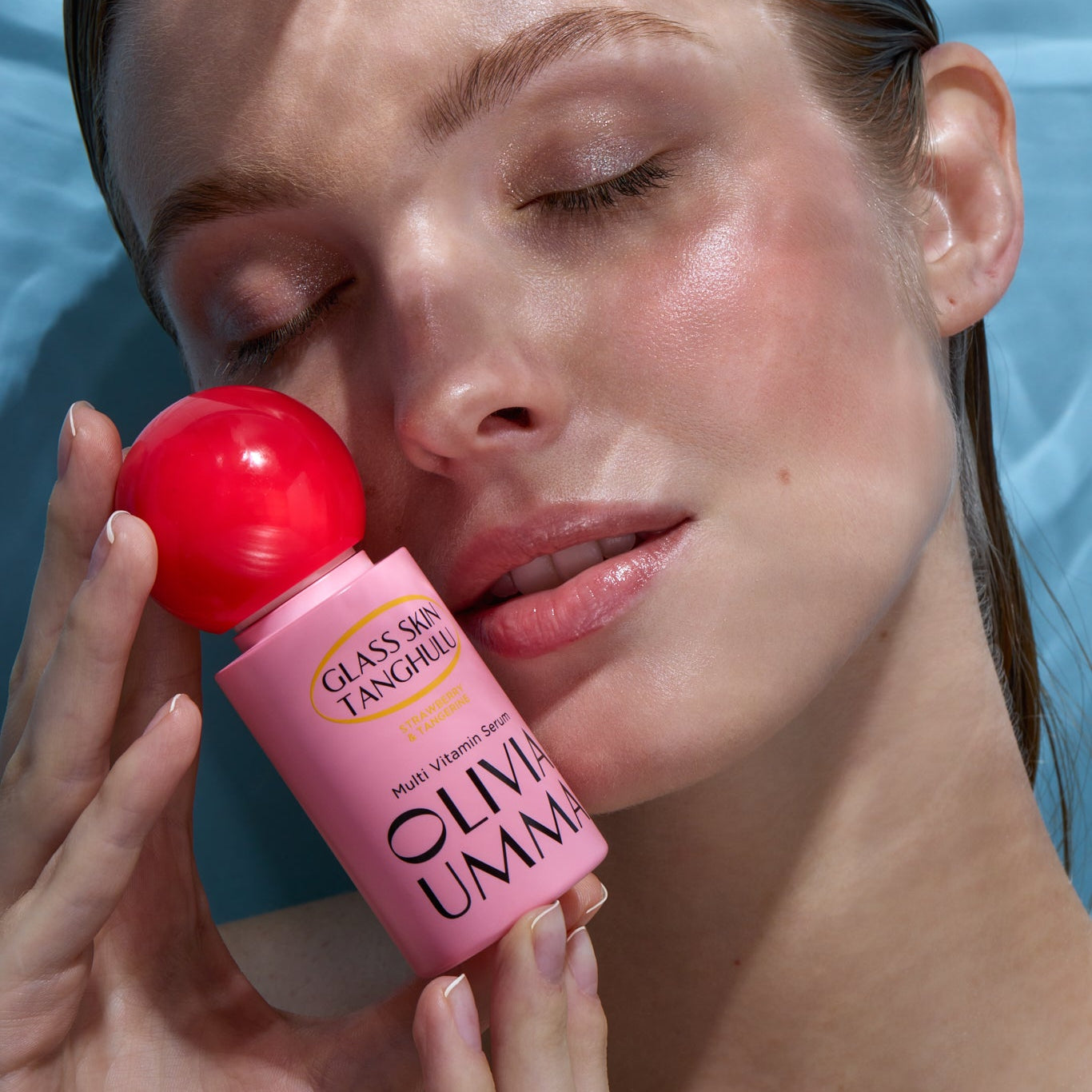 Woman applying olive oil to her face with a pink bottle labeled 'Olive Umma' against a blue background.