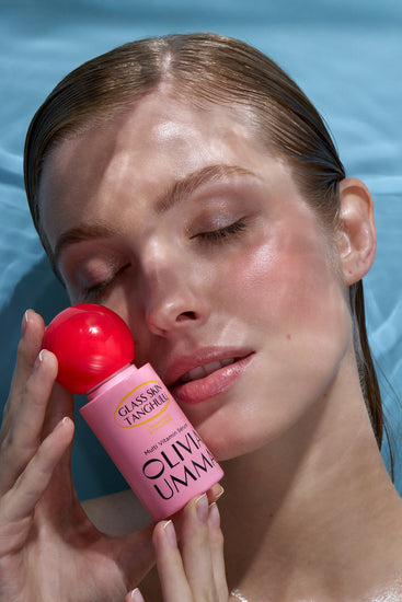 Woman applying olive oil to her face with a pink bottle labeled 'Olive Umma' against a blue background.